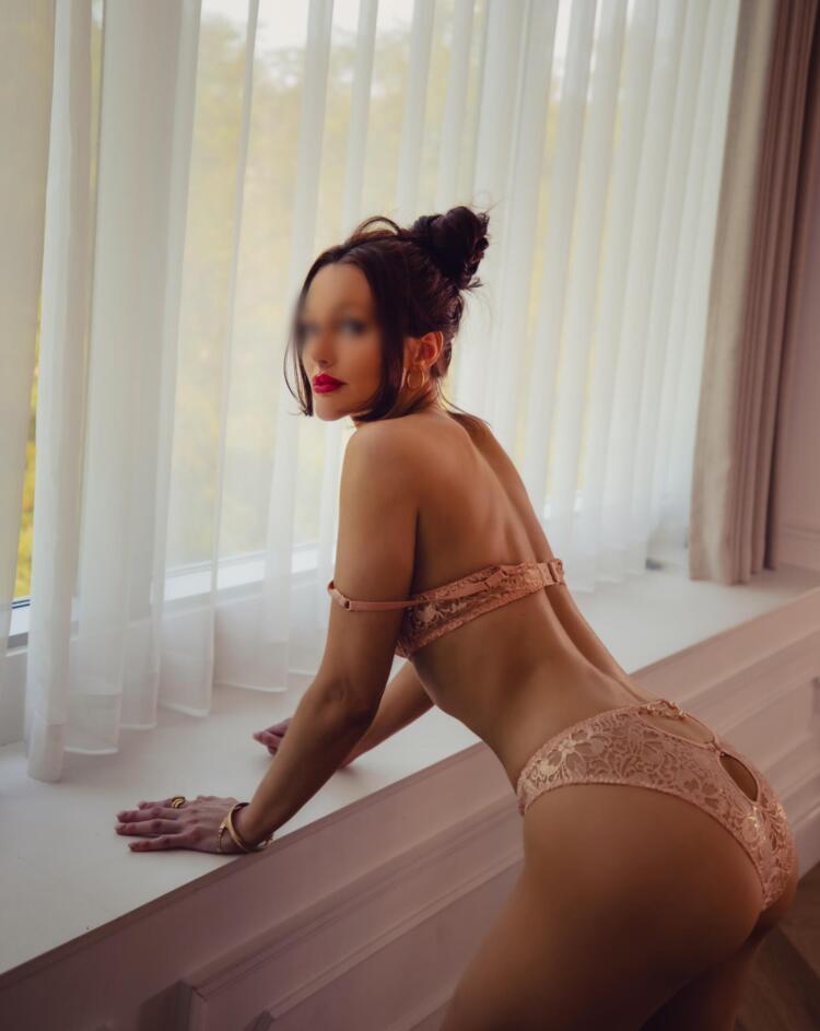 Escort with long brunette hair poses for the camera beside a window in pink lacey lingerie