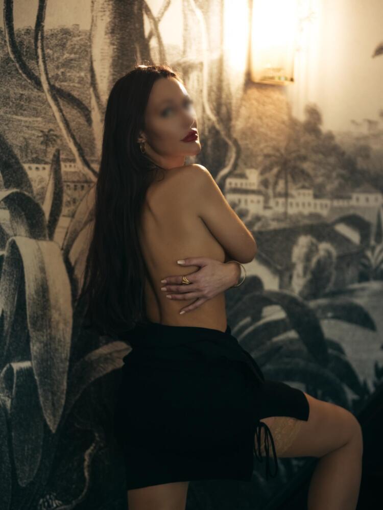 Elite travel companion looks back at you wearing thigh high stockings and a classic black dress, letting her long, brunette hair fall over her shoulders