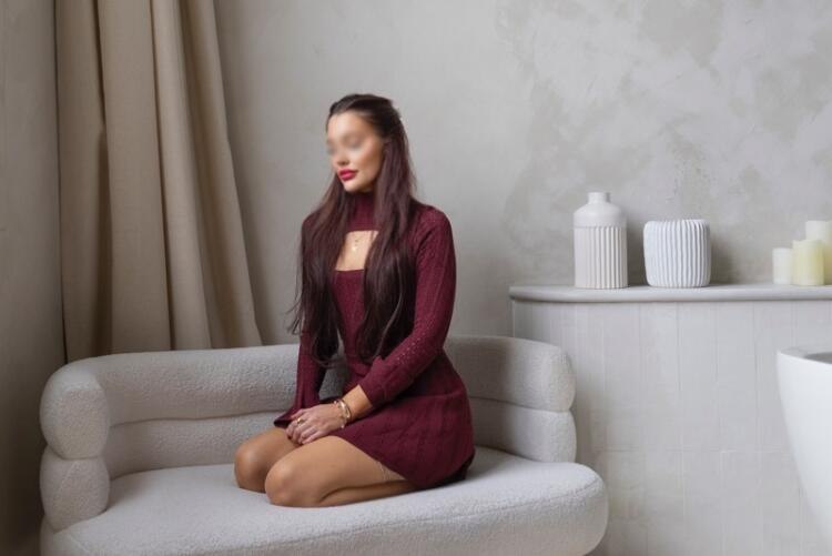 While sitting on a white pouf couch in Toronto, Mediterranean companion shows off her long brunette hair wearing a short red dress