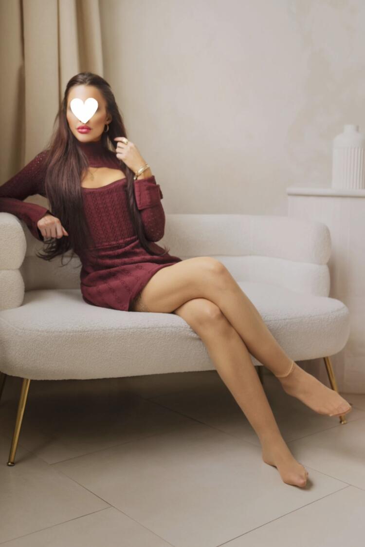 While sitting on a white pouf couch in Toronto, Mediterranean companion shows off her long legs and long brunette hair wearing a short red dress